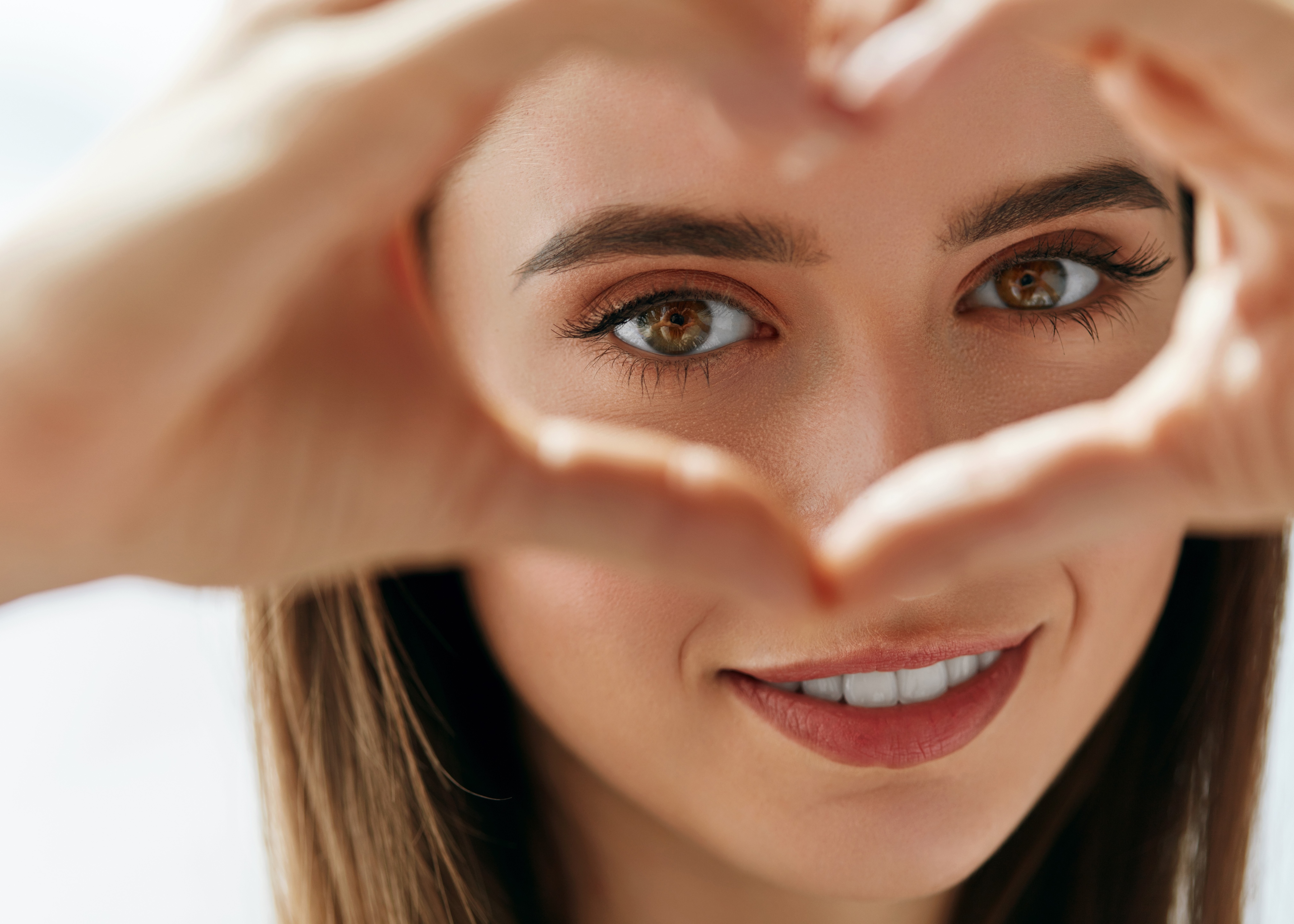 Close-up of a smiling woman forming a heart shape with her hands framing her eyes.