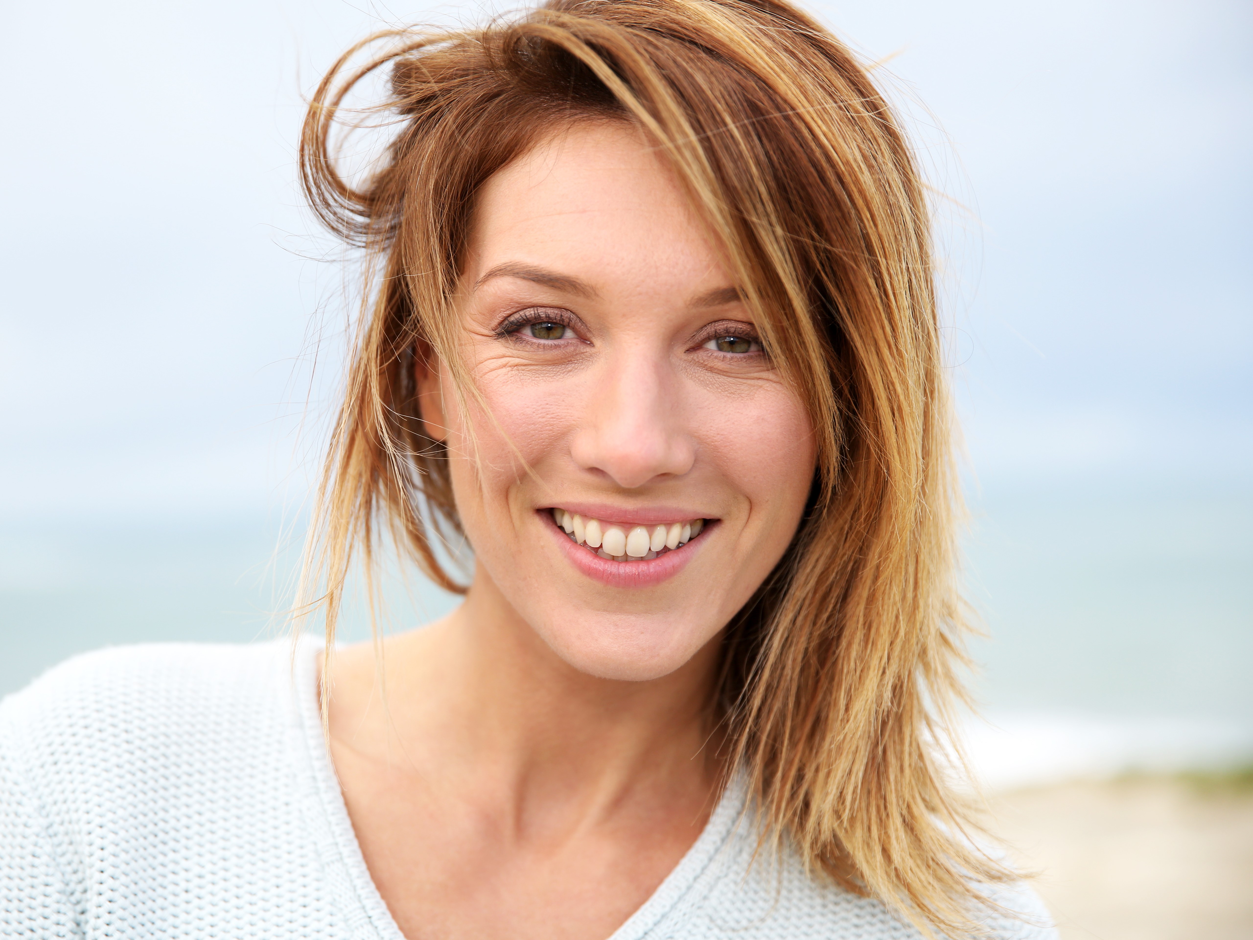 Close-up of a smiling woman with light brown hair wearing a light blue sweater outdoors.