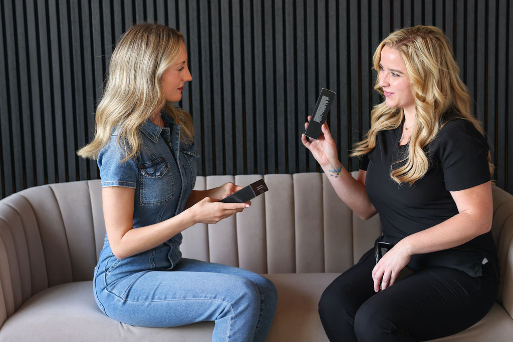 Two women sitting on a couch, discussing and holding black product boxes, against a black slatted wall background.