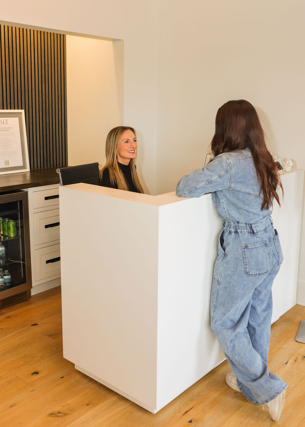 Receptionist smiling and talking to a woman in denim clothes leaning on a white reception desk in a modern office.