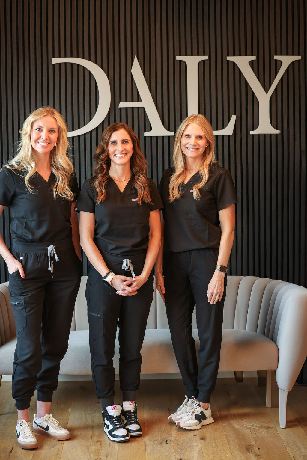 Three women in black scrubs standing and smiling in front of a black wall with large white letters spelling DALY.