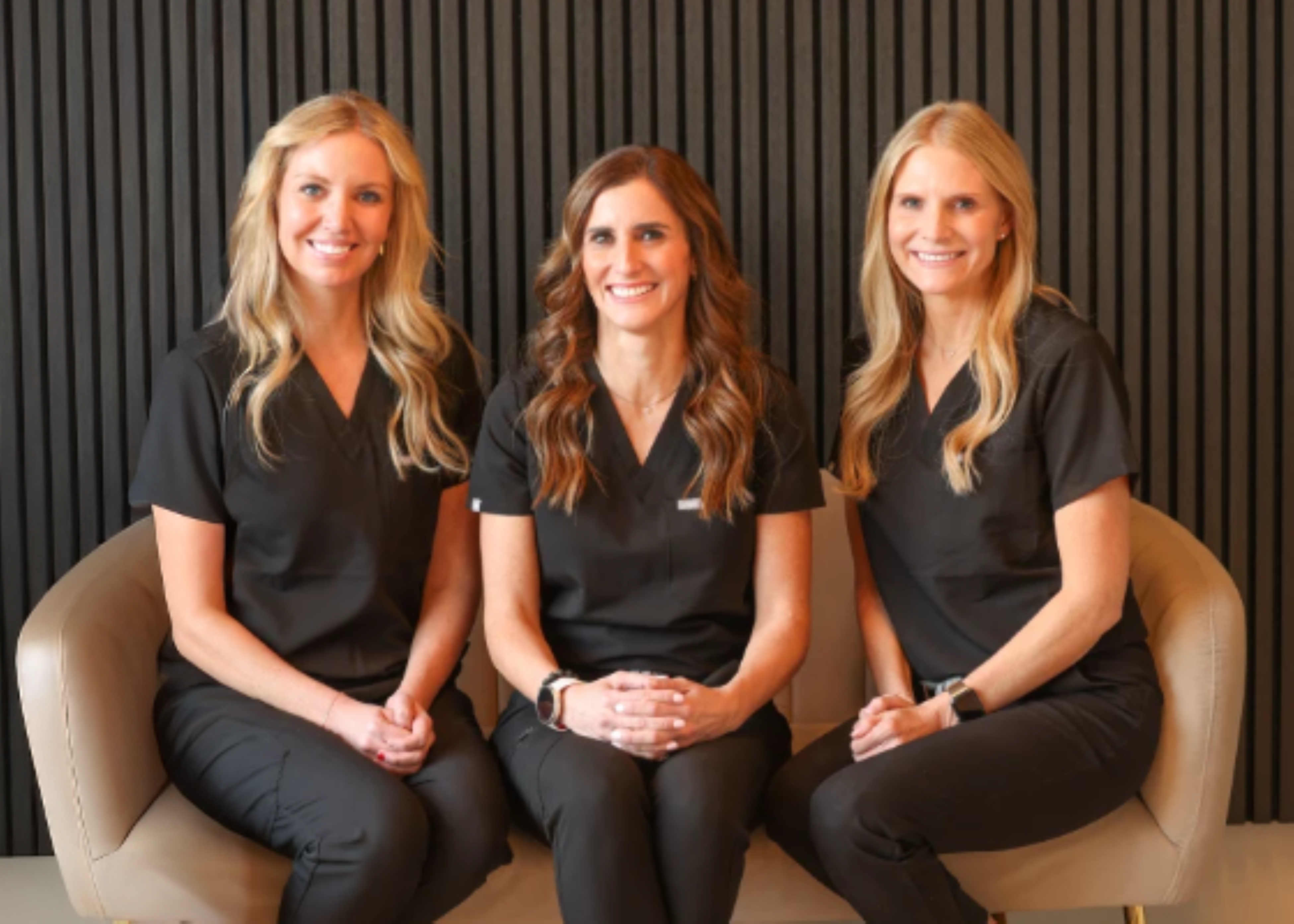 Three smiling women wearing black medical scrubs sitting side by side on beige chairs against a dark vertical panel background.