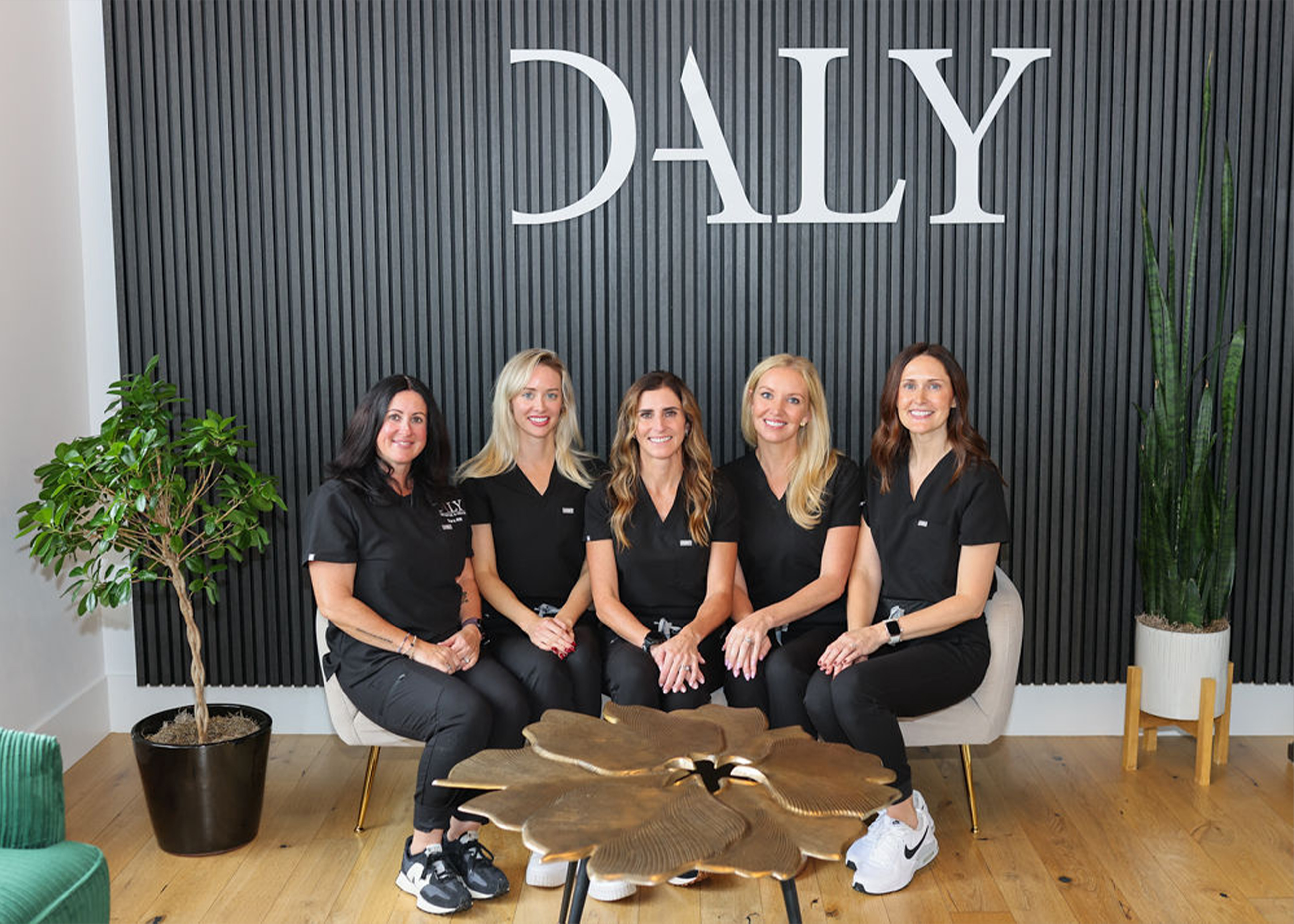 Five women wearing black uniforms sitting on chairs in front of a black vertical panel wall with the word 'DALY' above them and a gold leaf-shaped table in front.