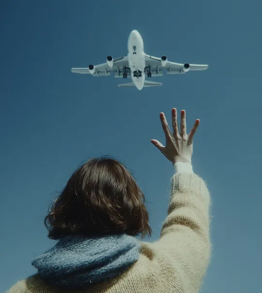 Person with short hair wearing a beige sweater and blue scarf reaching up towards a low flying airplane against a clear blue sky.