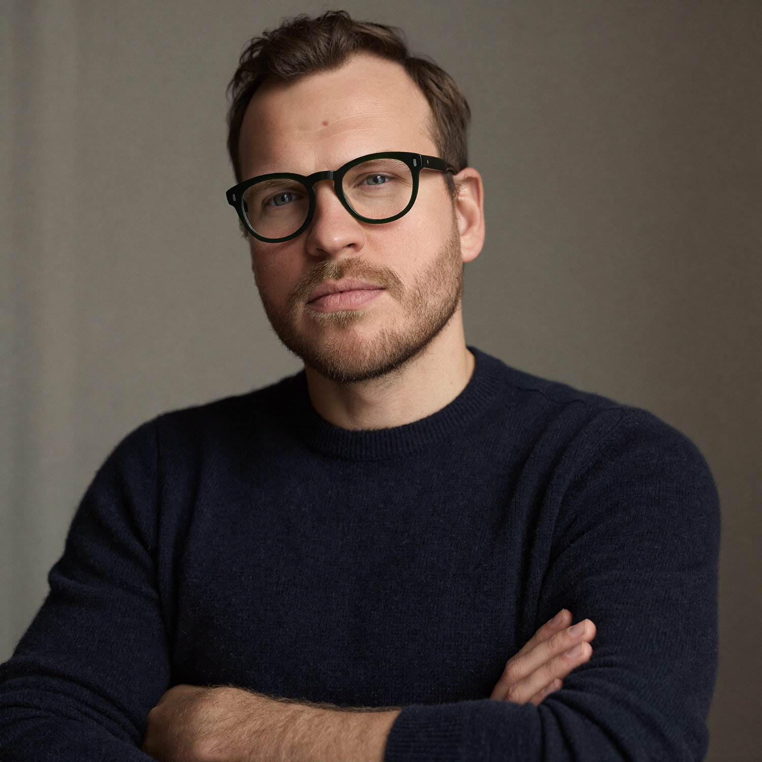 Man with wavy brown hair and beard wearing round glasses and a brown sweater over a white shirt in warm light.