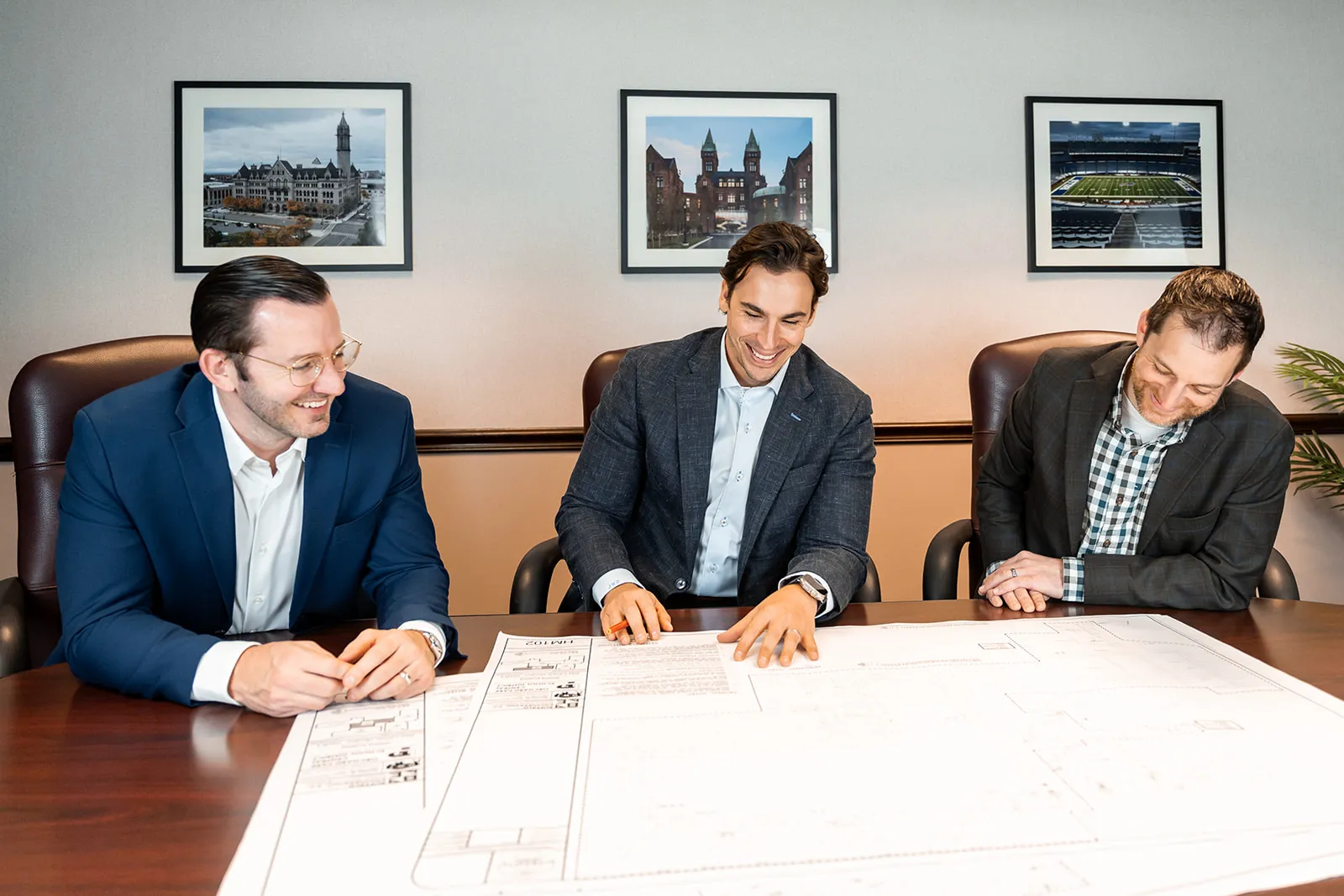 Three men in business attire sitting around a table, reviewing large architectural plans, smiling and discussing.