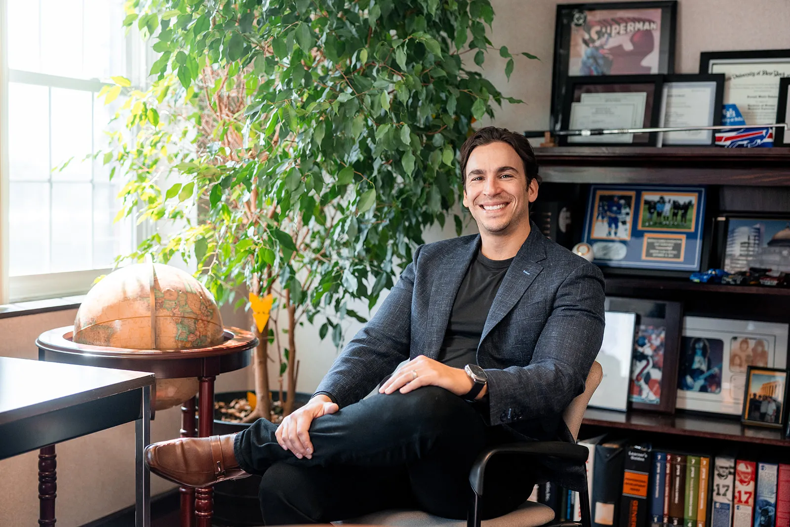 Smiling man in a blazer sitting cross-legged on a chair in an office with a globe, large plant, and shelves with framed certificates and photos.