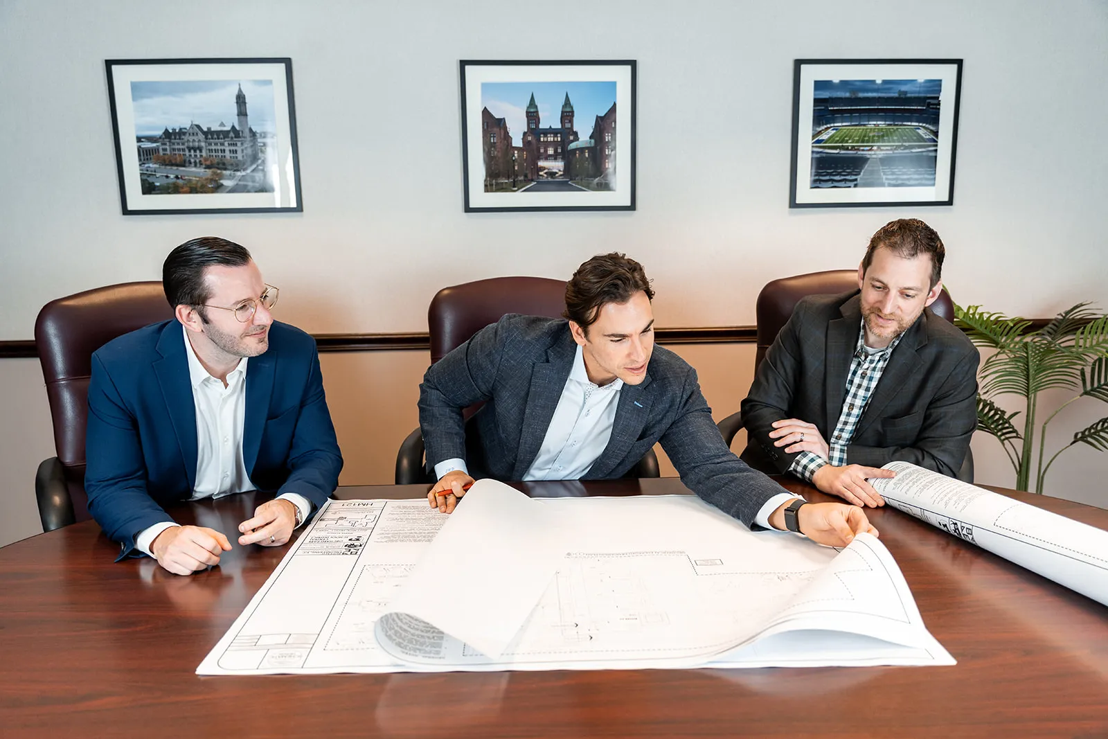 Three men in business attire sitting at a large wooden table reviewing architectural blueprints.