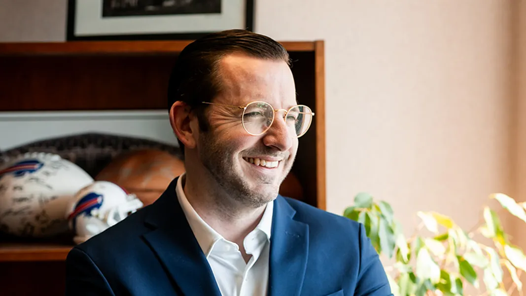 Man with glasses wearing a blue suit and white shirt smiling indoors near a bookshelf with sports helmets and a plant.