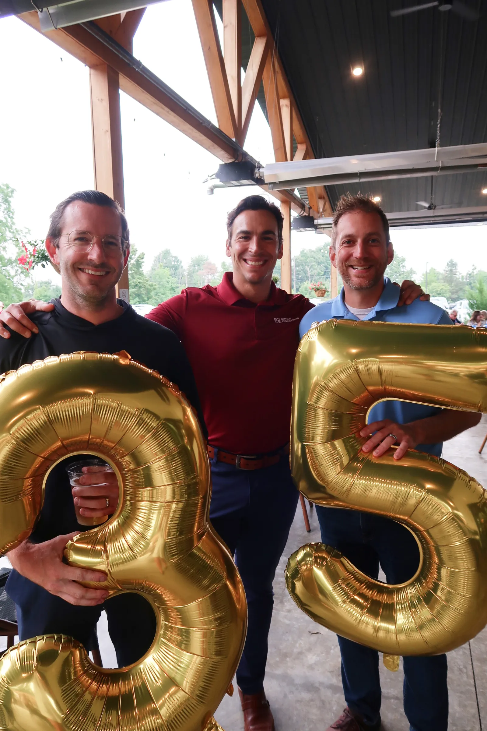 Three men standing under a covered outdoor area, smiling, holding large gold balloons shaped like the numbers 3 and 5.