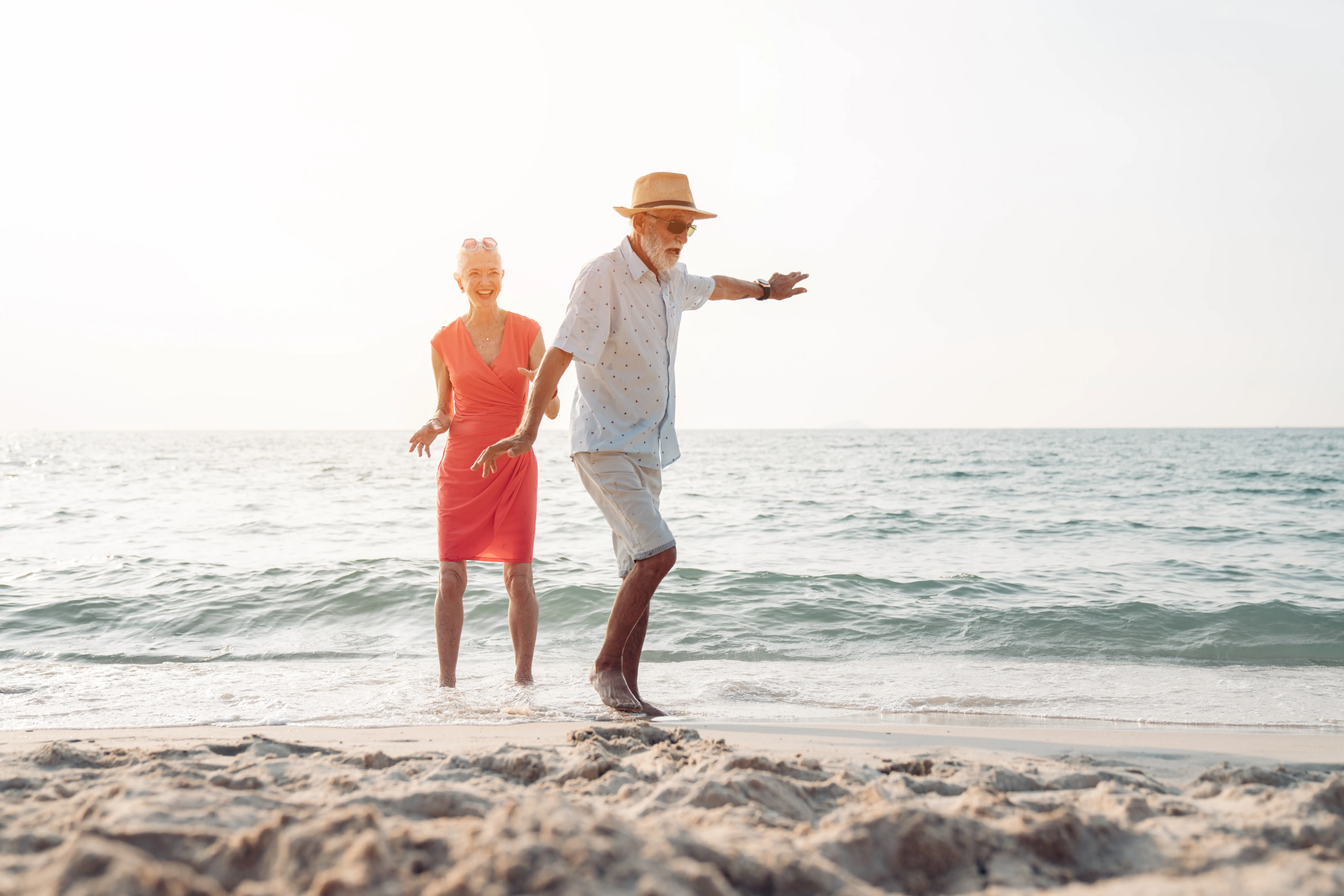 Elderly couple enjoying a sunny day at the beach with the man balancing on one foot in the shallow water.