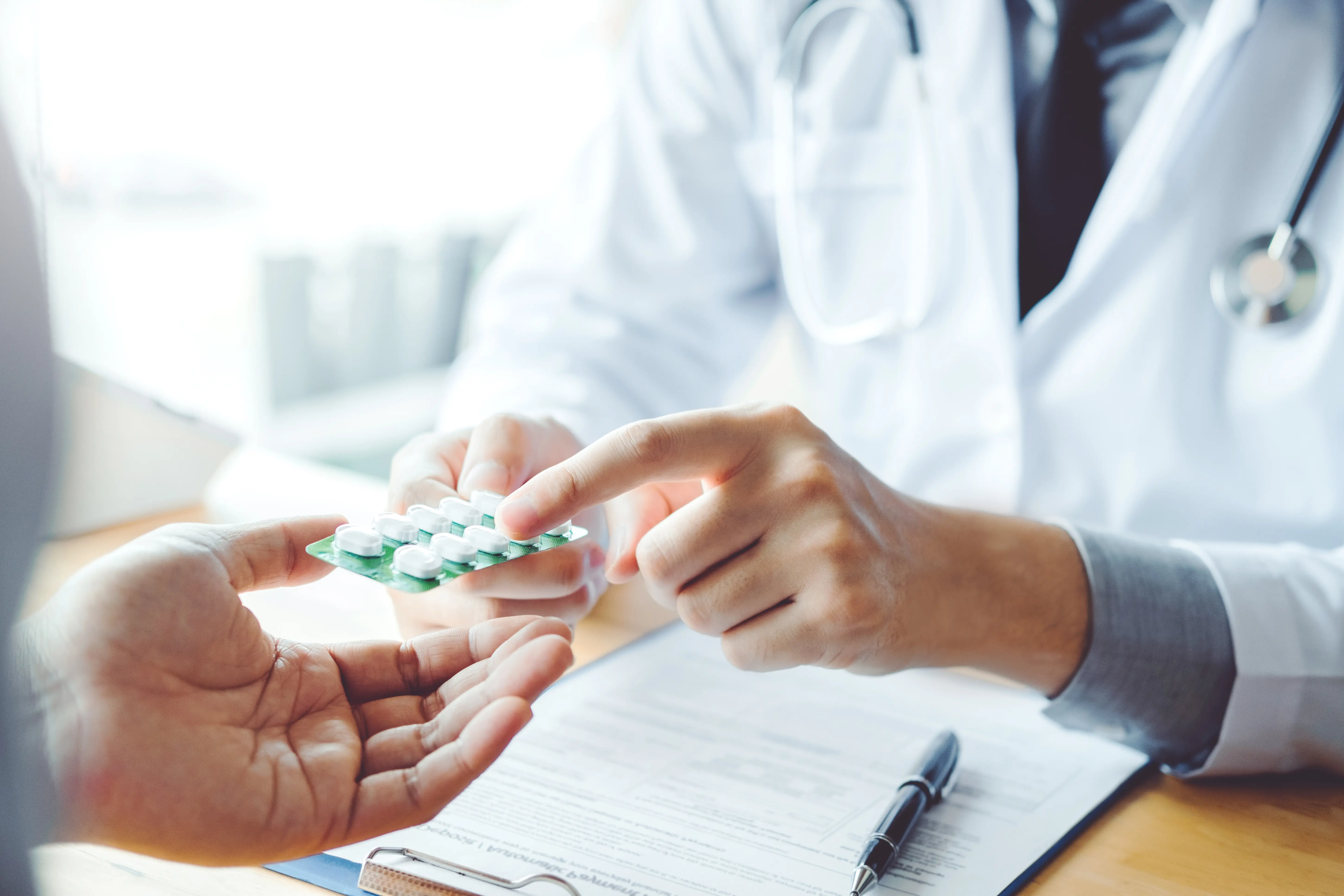 Doctor handing a blister pack of white pills to a patient over a table with medical documents and pen.