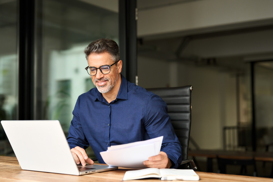 Man wearing glasses and a blue shirt sitting at a wooden desk, smiling while working on a laptop and holding papers, with a notebook open on the desk.
