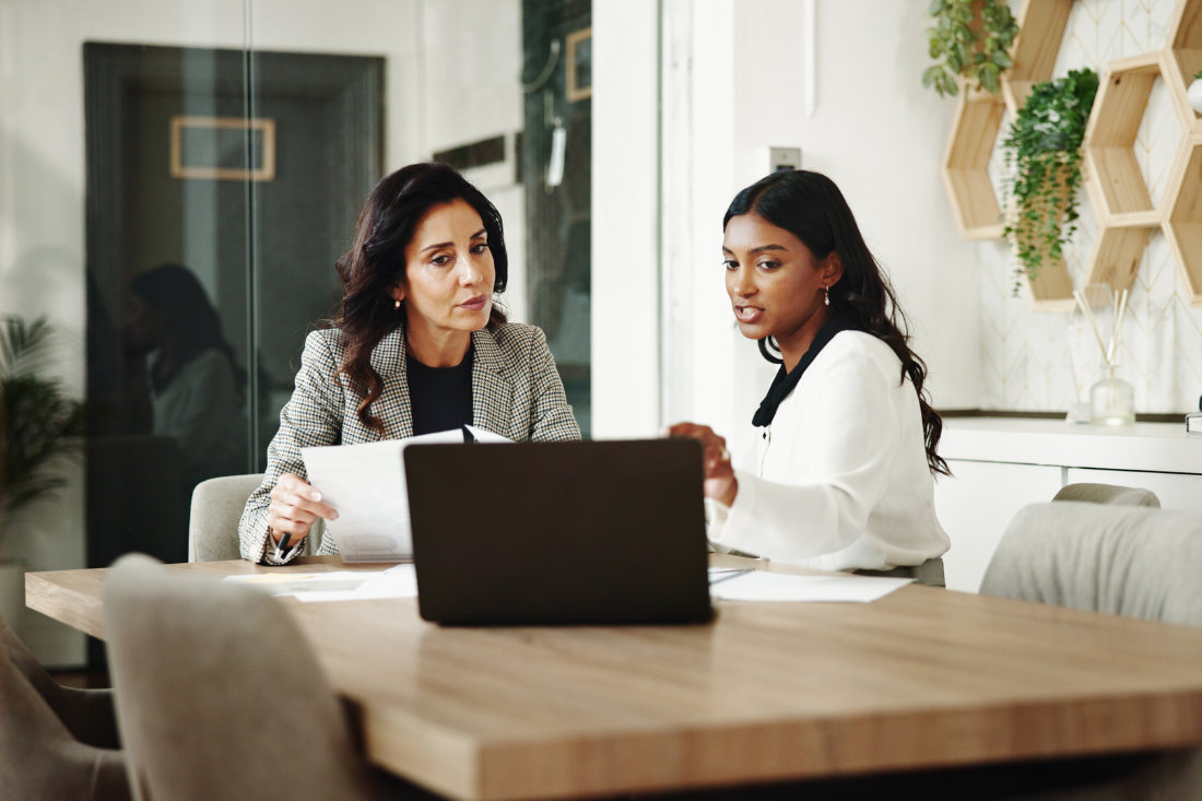 Two professional women seated at a wooden table in an office discussing documents while looking at a laptop screen, with wooden hexagonal shelves and green plants on the wall behind them.