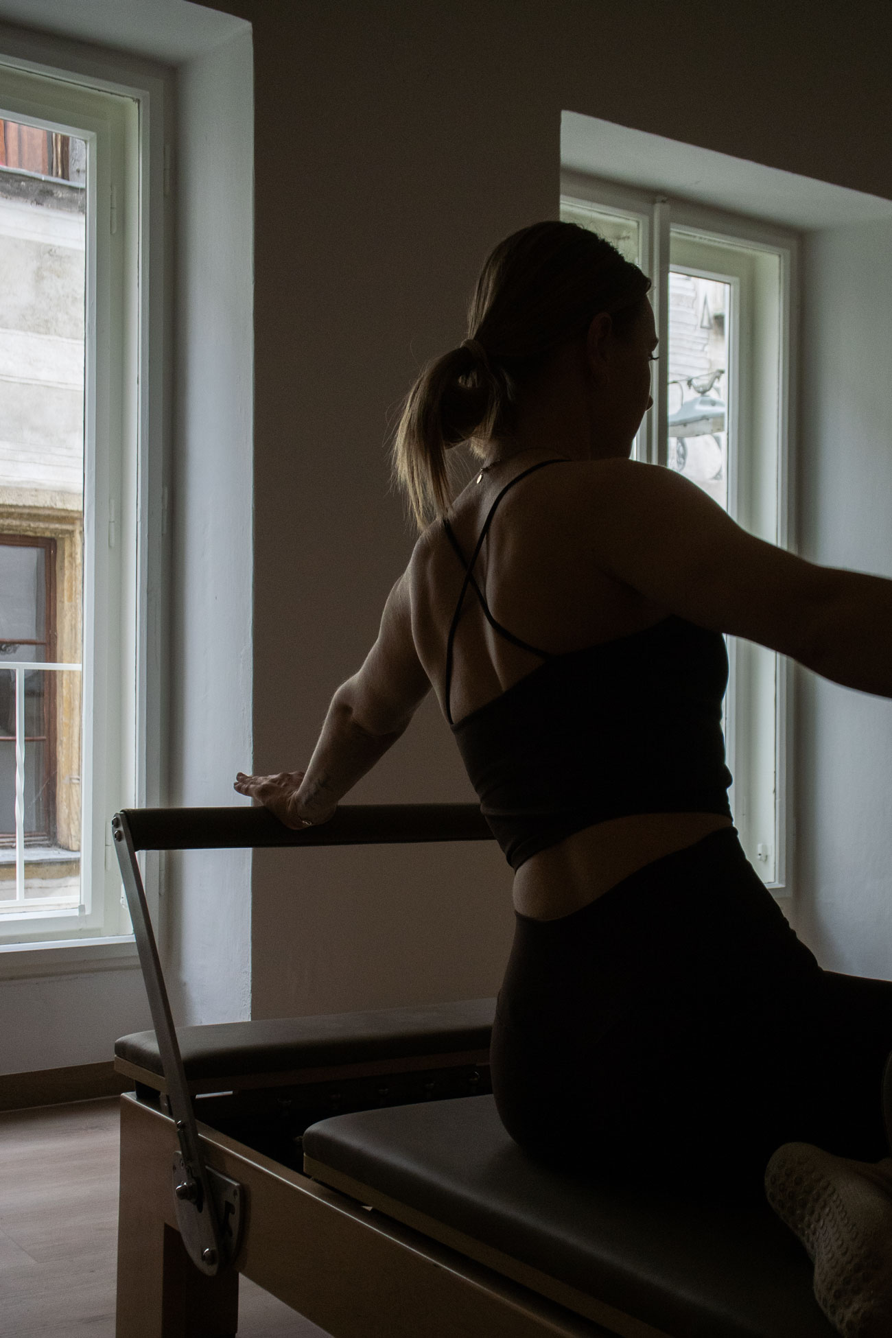 Woman holding a Pilates reformer bar in a dimly lit studio with large windows.