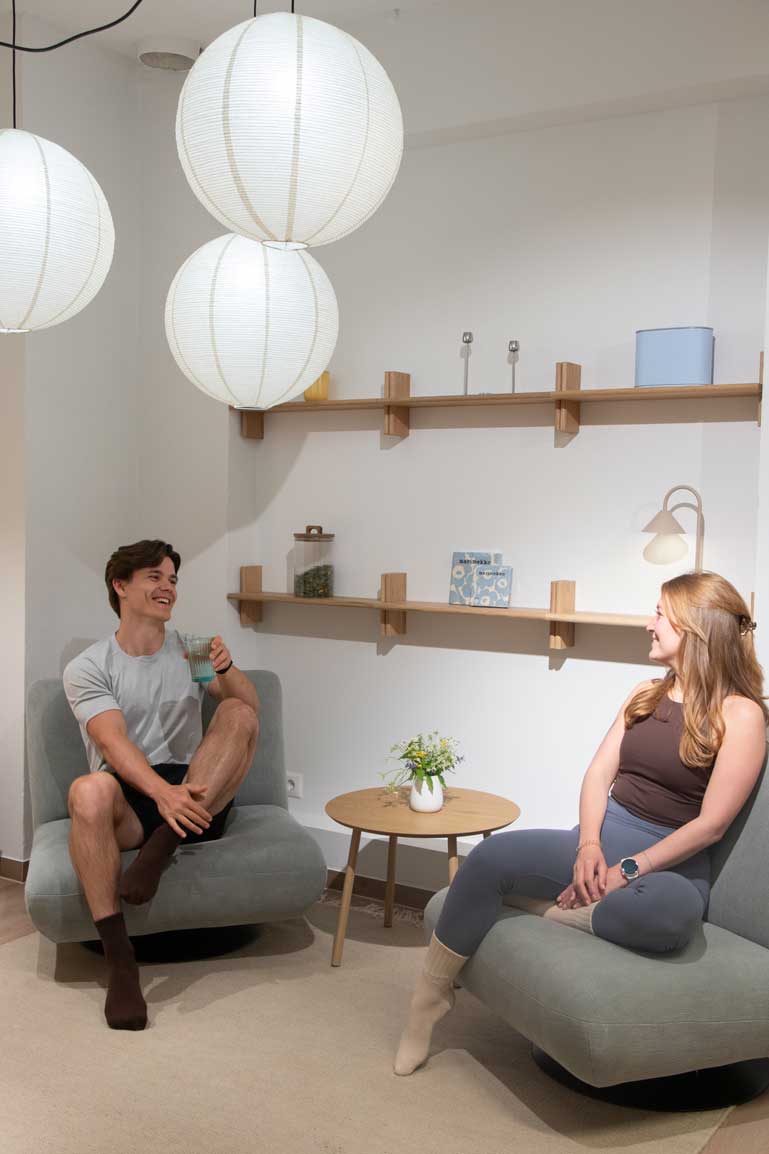 Man and woman sitting on gray chairs in a room with round hanging lamps and wooden shelves.