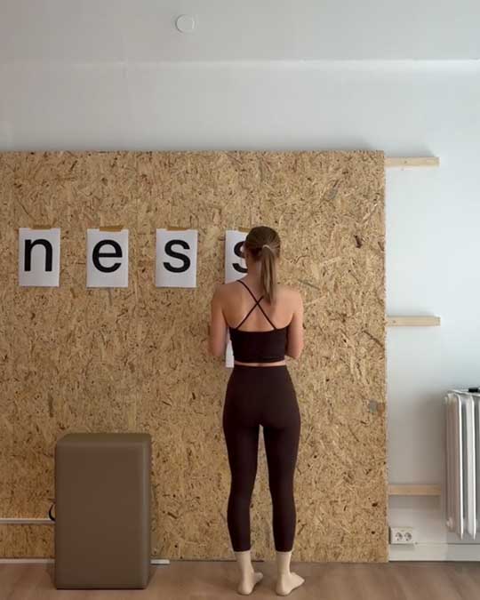 Woman in black workout clothes standing barefoot in front of wall with letters 'n', 'e', 's', 's'.