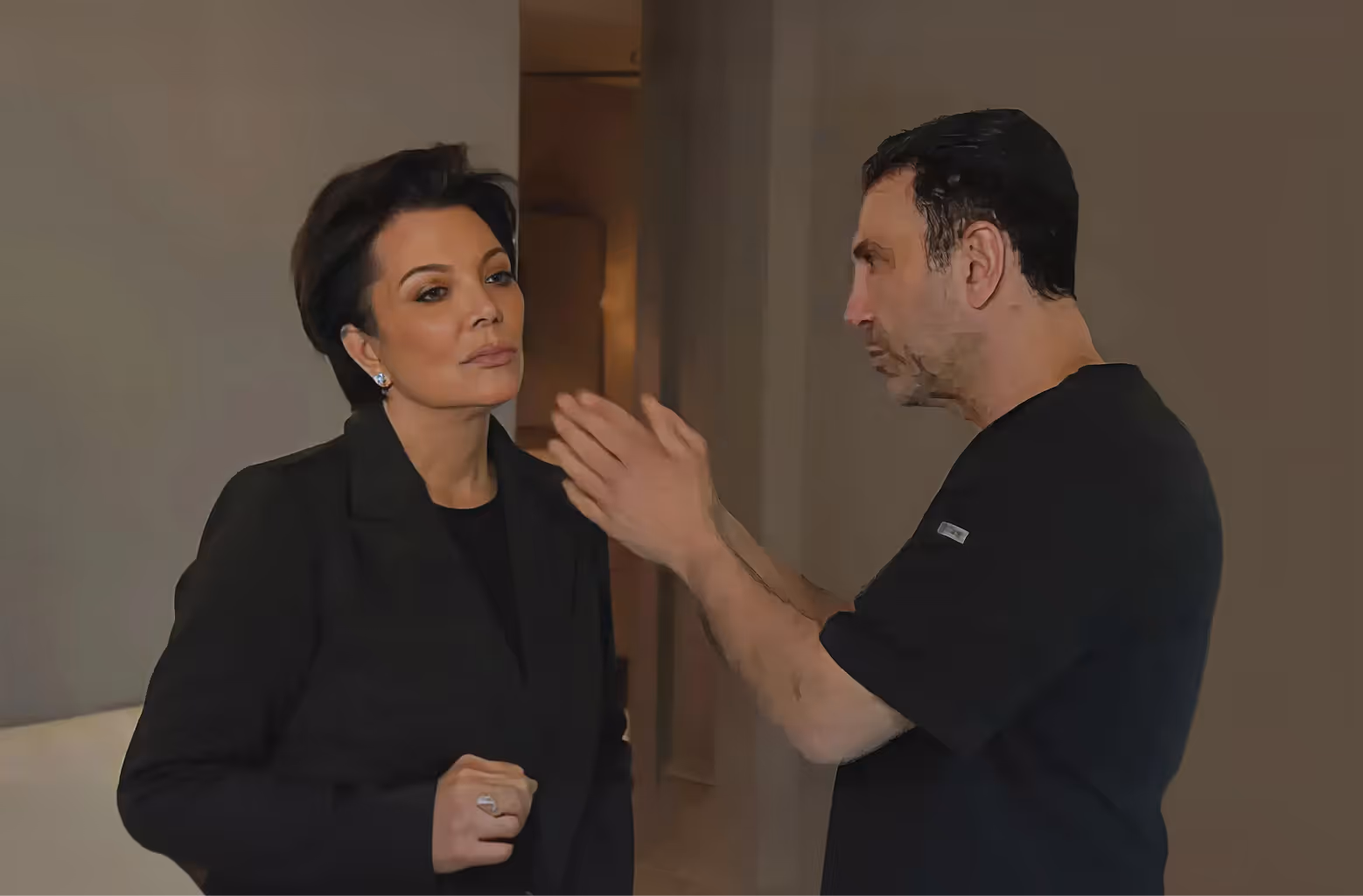 A woman in a black blazer listens thoughtfully while a man in a black shirt gestures and speaks to her indoors.