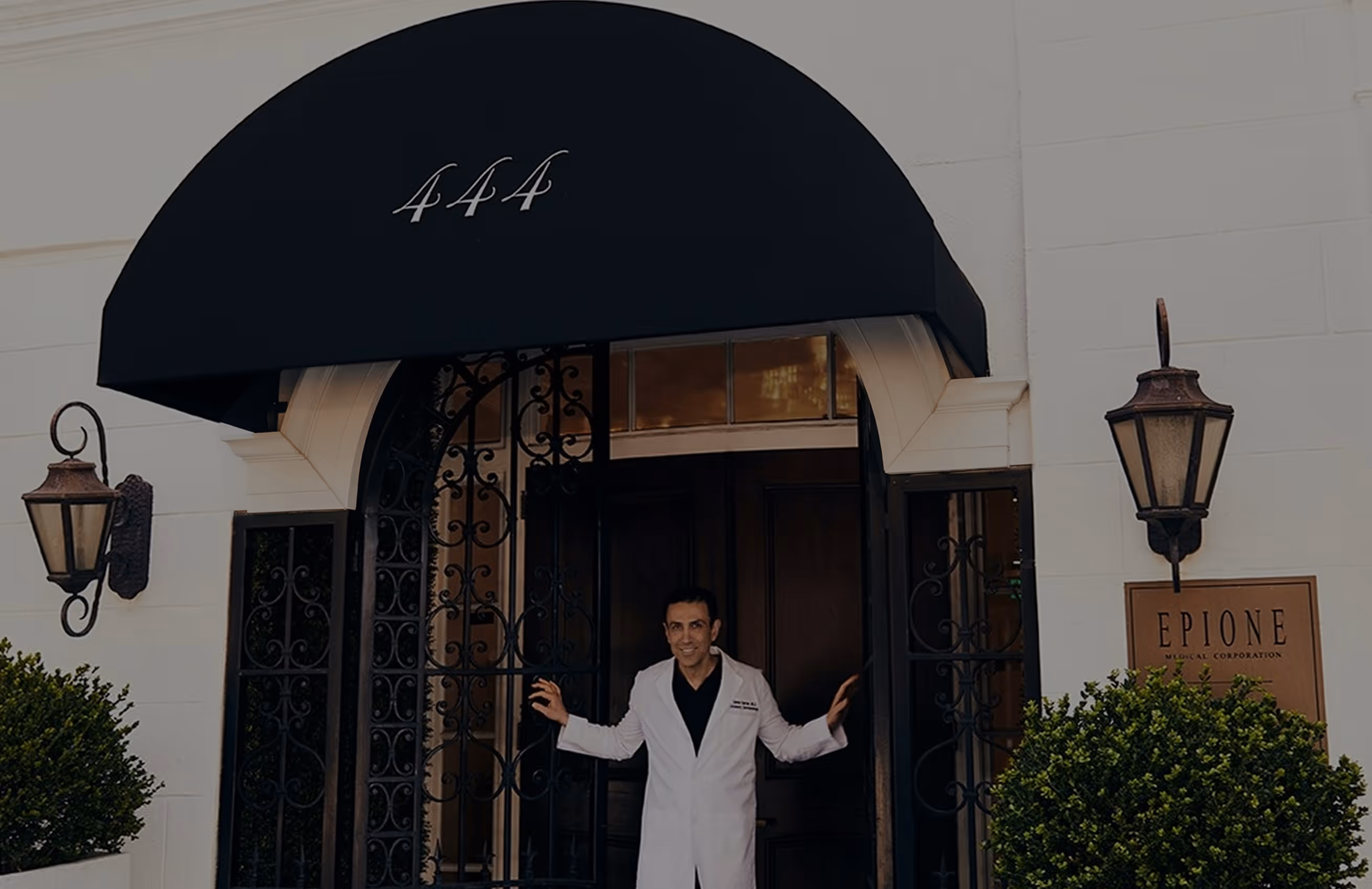 Man in white coat standing in the doorway under a black awning with the number 444 above.