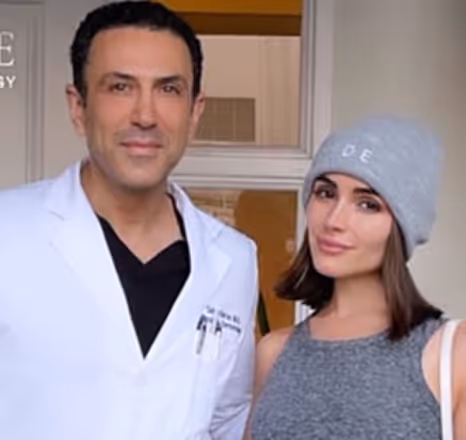 Man in a white medical coat standing next to a woman wearing a gray sleeveless top and a gray beanie.