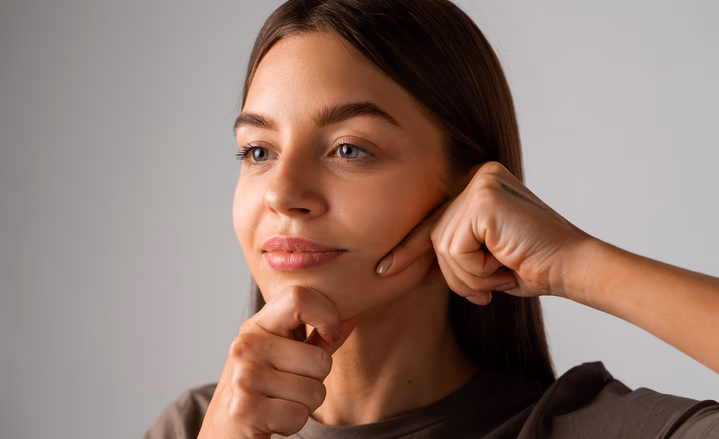 Young woman using fists to massage her cheeks against a plain background.