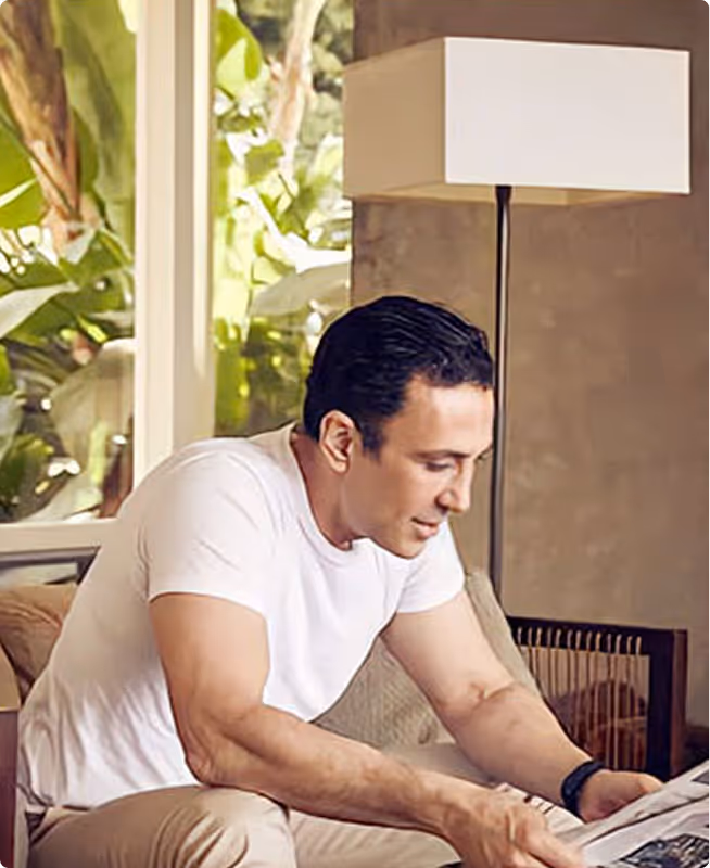 Man in a white t-shirt sitting on a couch, reading a newspaper indoors near a window with green plants outside.
