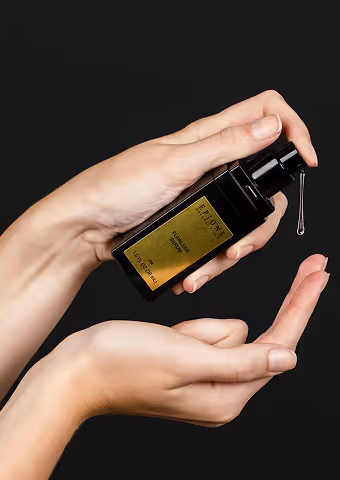 Hands holding a black dropper bottle with a golden label dispensing a drop onto a fingertip against a black background.