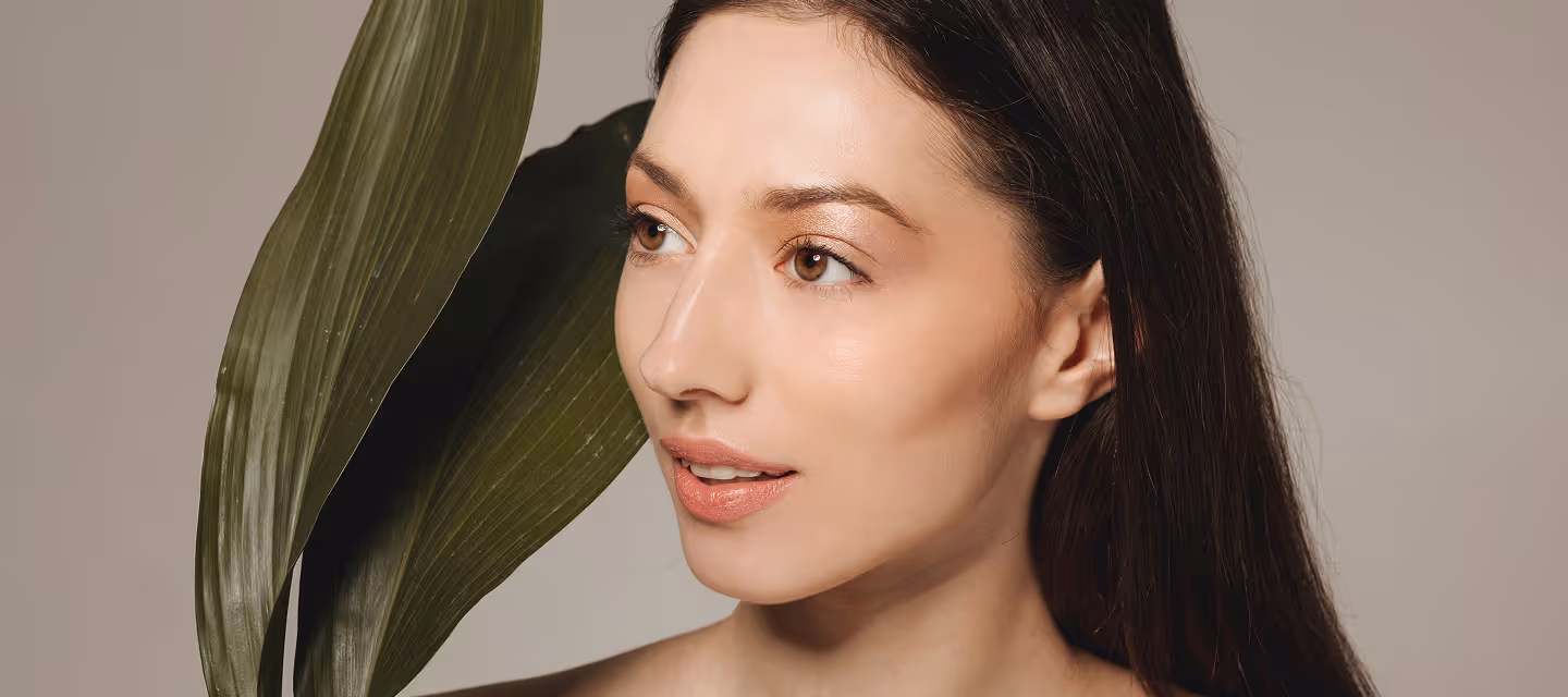 Close-up side view of a woman with long dark hair looking to the left, beside two large green leaves.