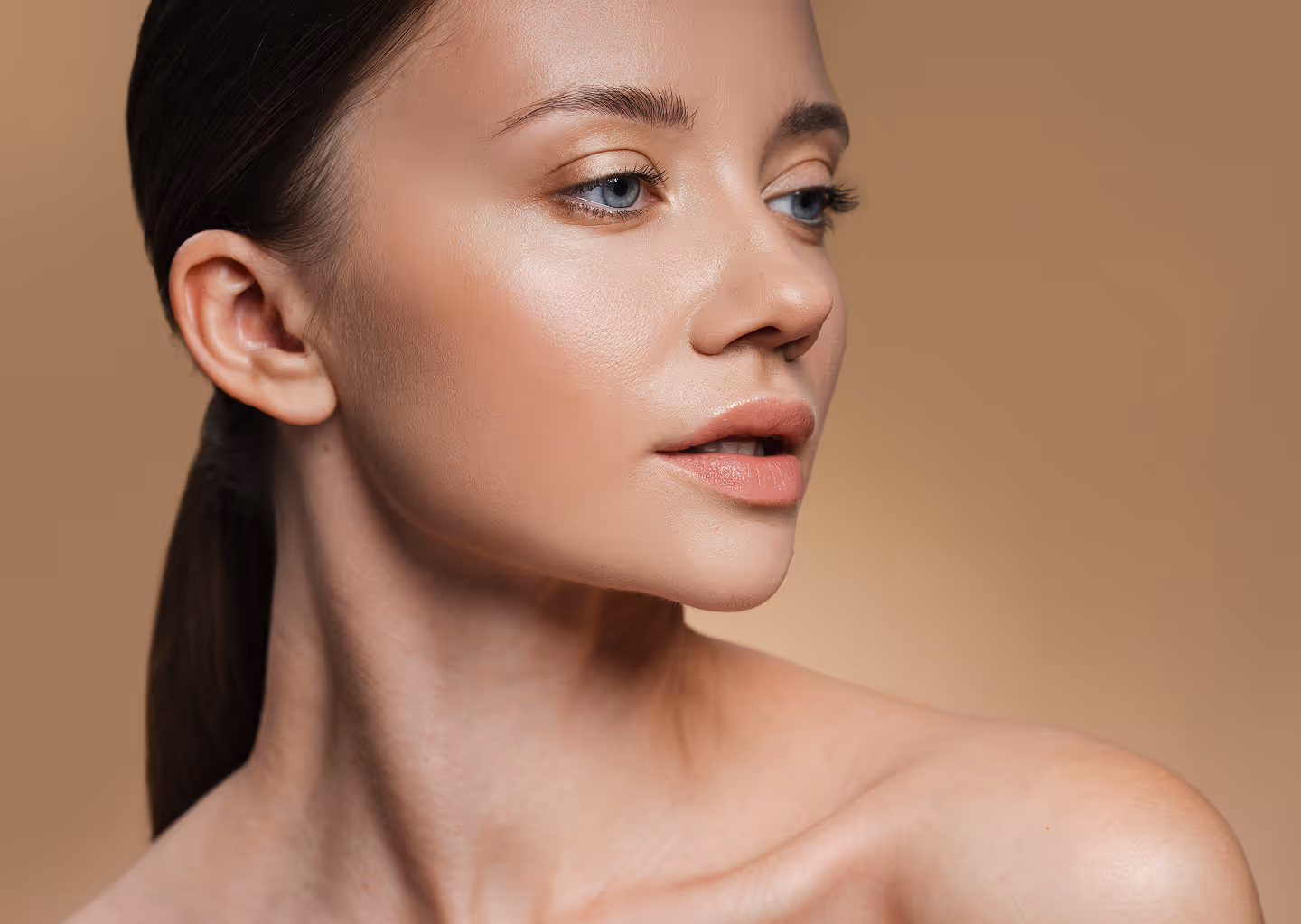 Close-up side profile of a woman with clear skin and blue eyes looking to the right against a beige background.