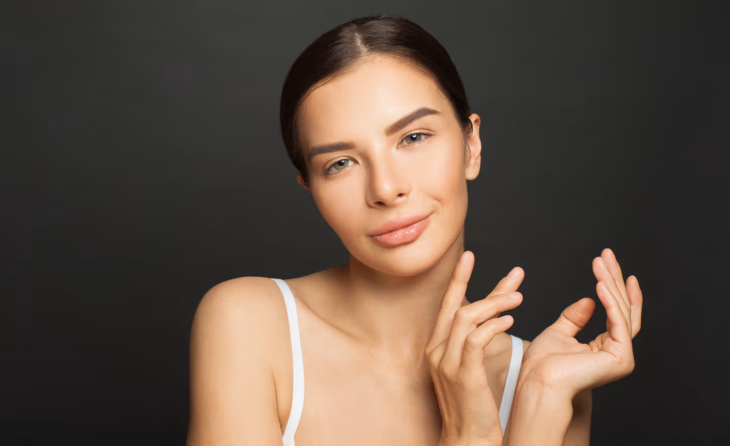 Young woman with clear skin and dark hair pulled back, softly smiling with hands raised near her face against a dark background.
