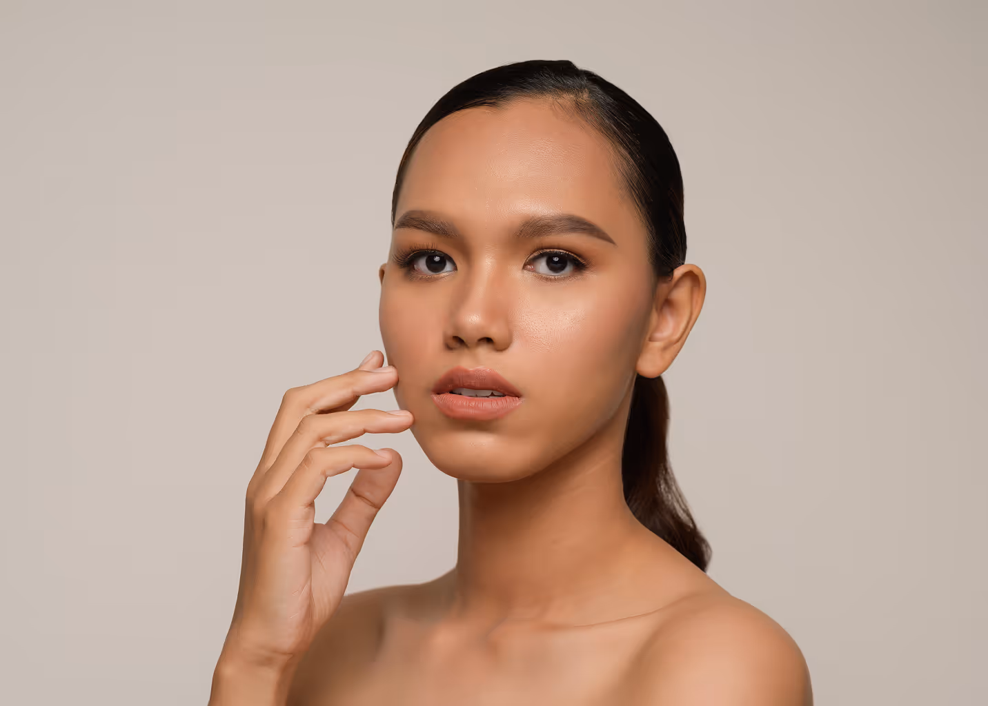 Close-up portrait of a woman with smooth skin and natural makeup touching her face against a neutral background.