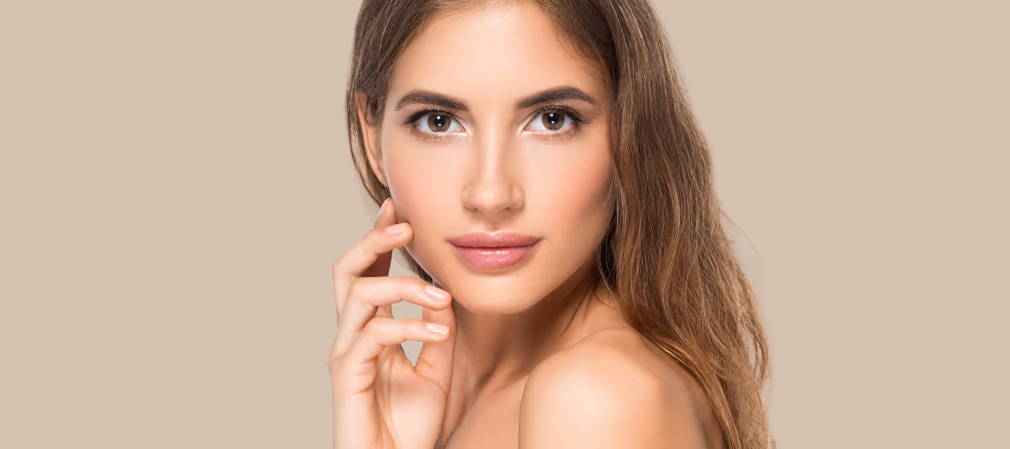 Close-up of a woman with long brown hair and natural makeup touching her face against a beige background.