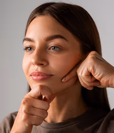Young woman gently pinching her cheek with both hands while looking slightly to the side.