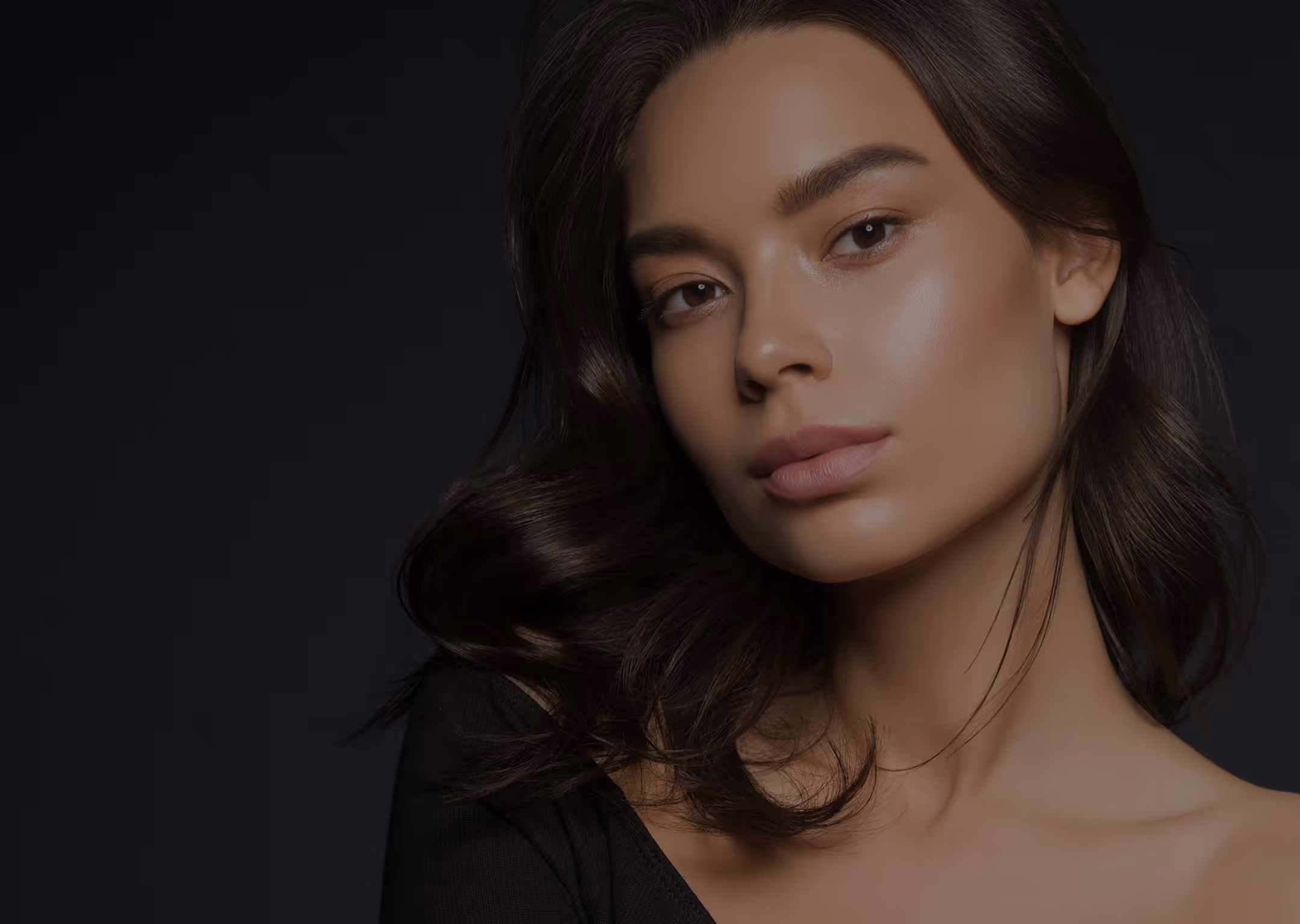 Close-up of a woman with smooth skin, dark hair, and neutral makeup against a dark background.