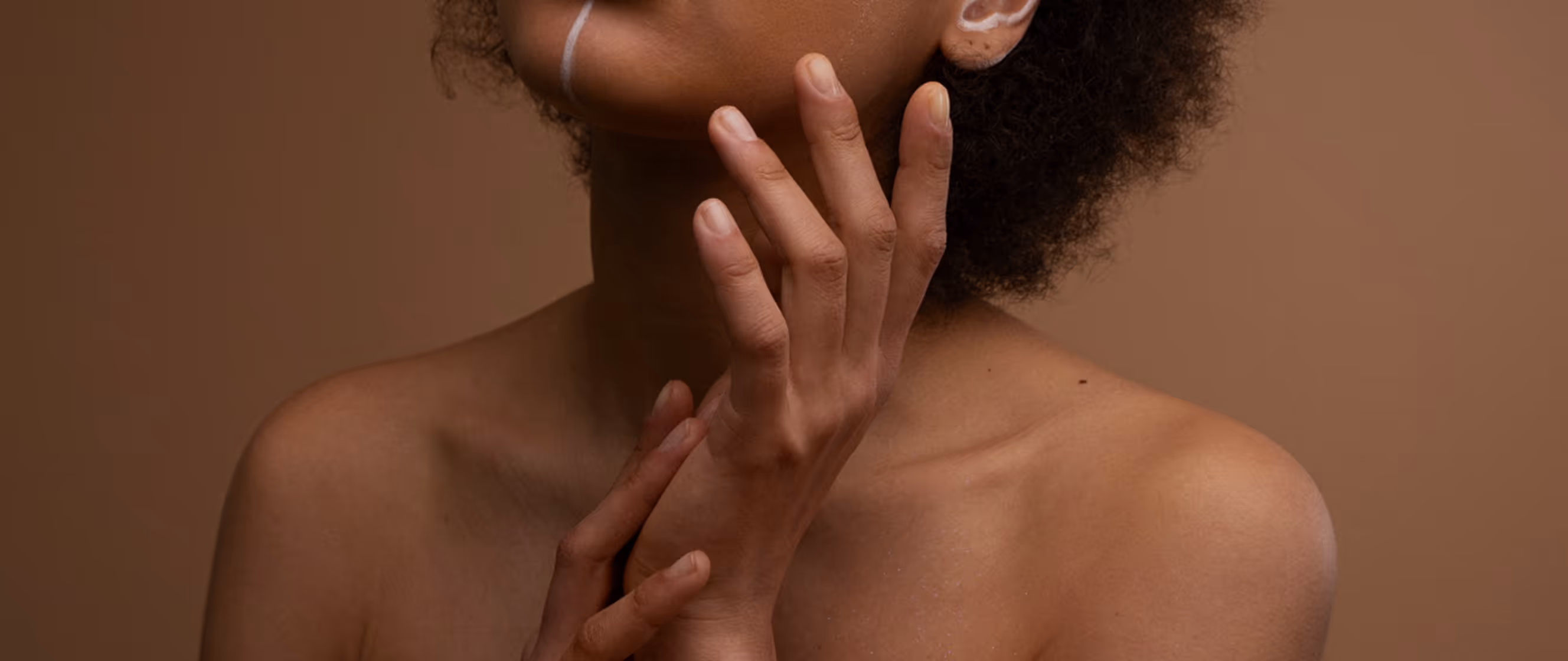 Close-up of a bare-shouldered person touching their neck and hand with a brown background.