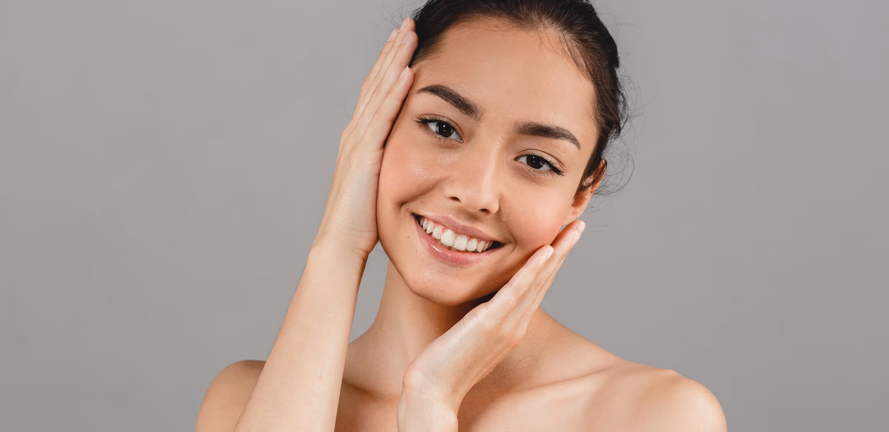 Smiling woman with clear skin touching her face gently on a gray background.