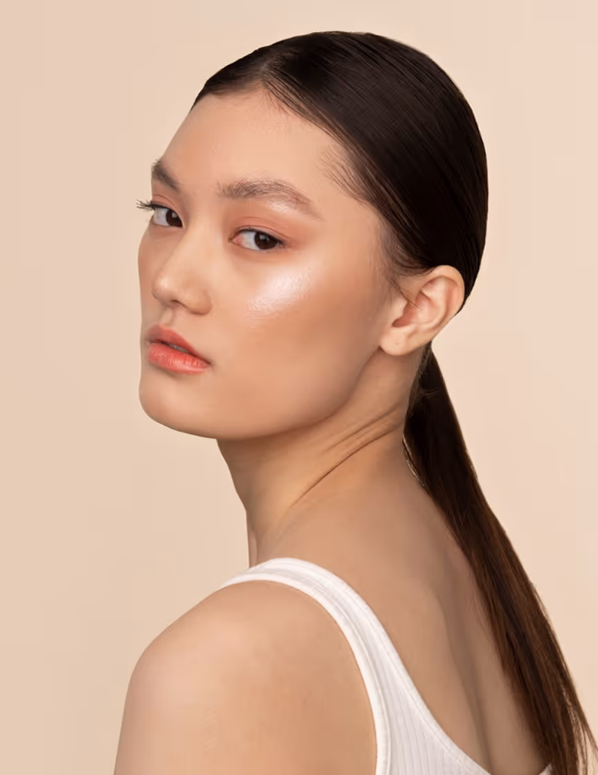 Young woman with smooth skin, tied-back dark hair, and subtle makeup against beige background.