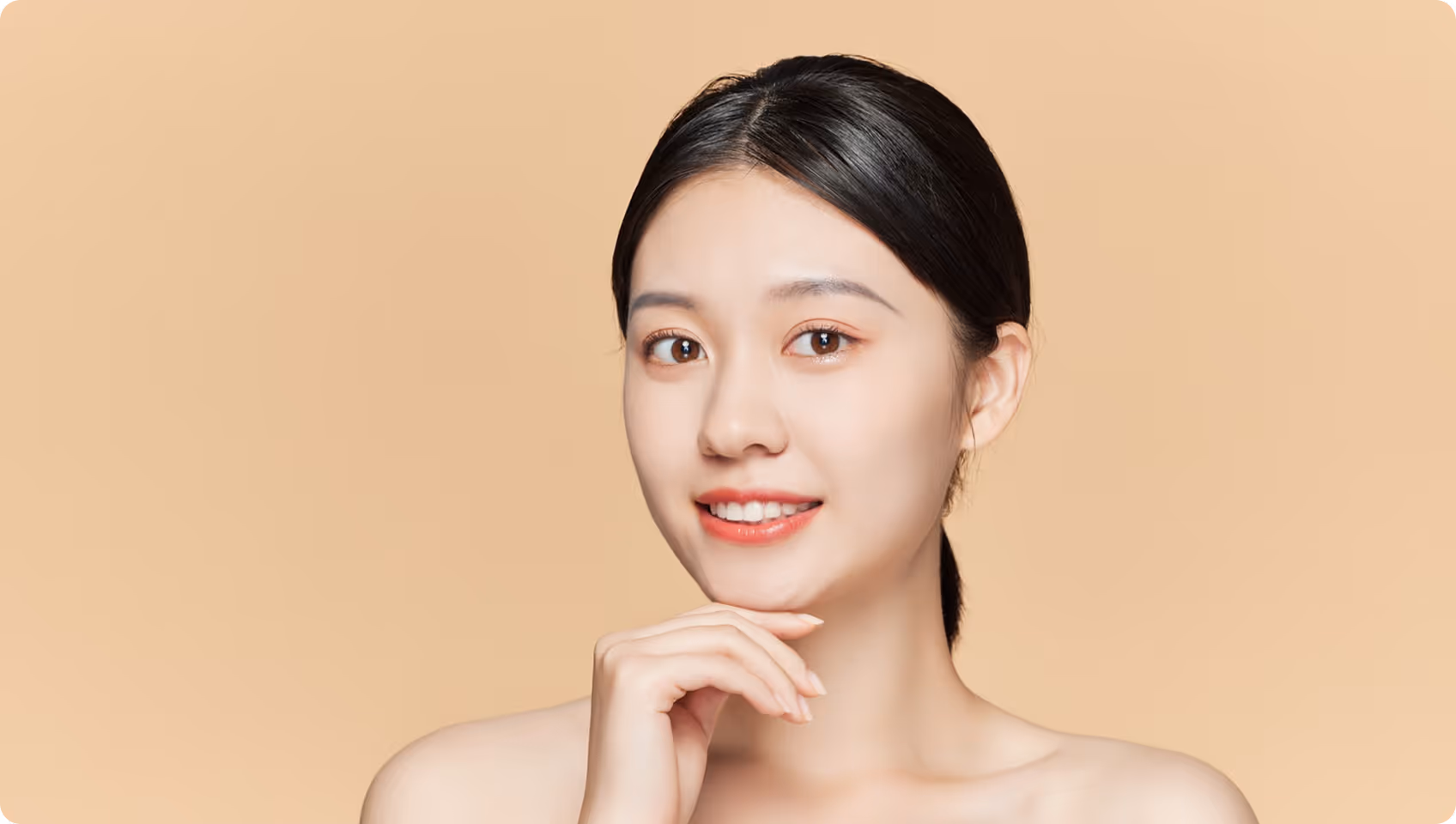 Young woman with clear skin smiling, resting chin on hand against beige background.
