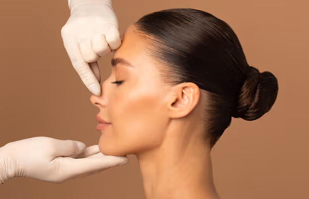 Woman with eyes closed having her nose examined by gloved hands against brown background.