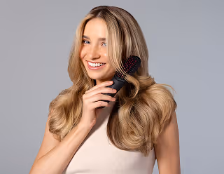 Smiling woman brushing her long blonde hair with a round hairbrush against a gray background.