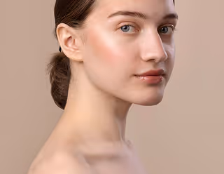 Young woman with smooth skin and hair tied back looking at camera against beige background.