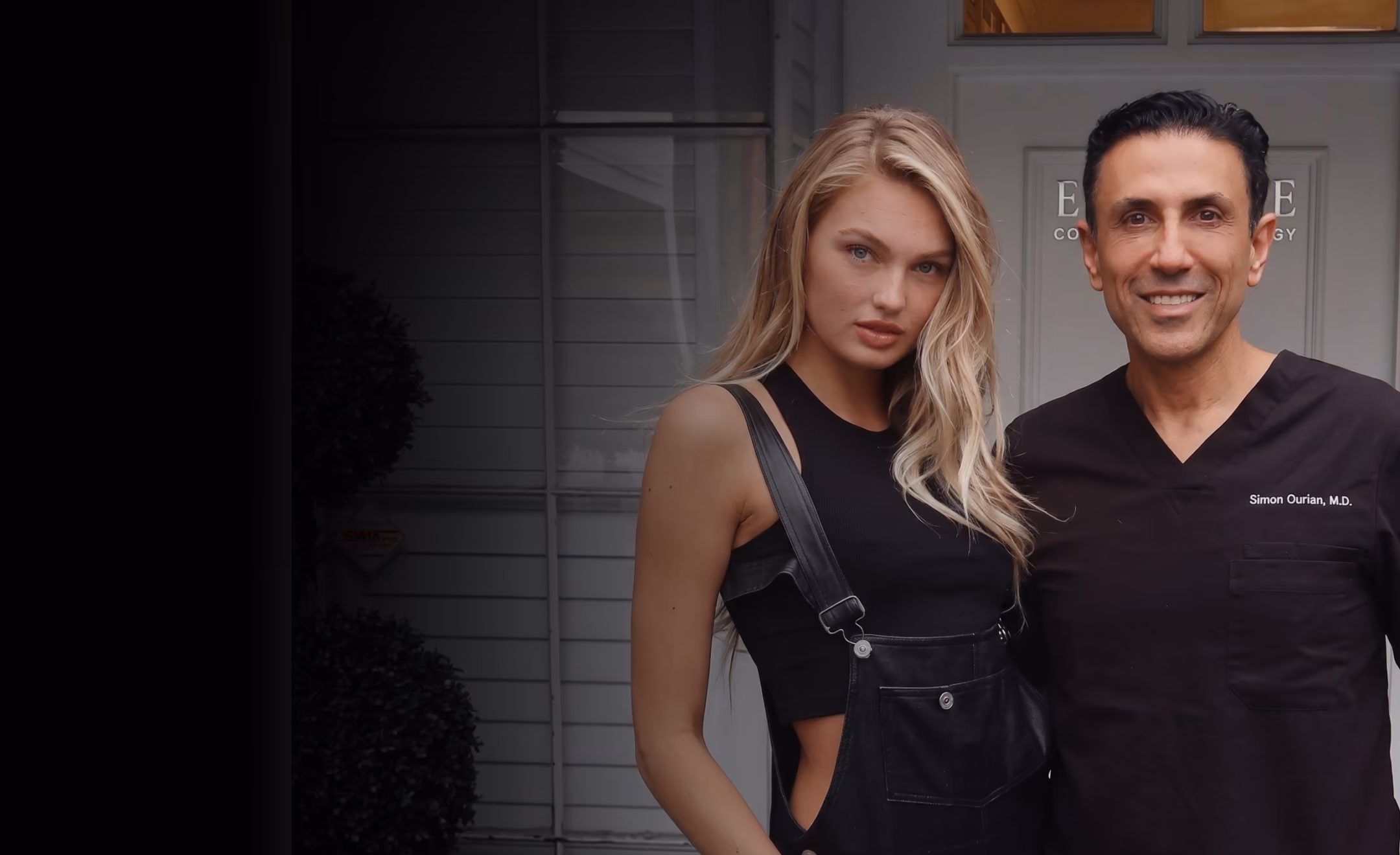 Smiling man in medical scrubs with a woman in black sleeveless top and overalls standing outside a building.