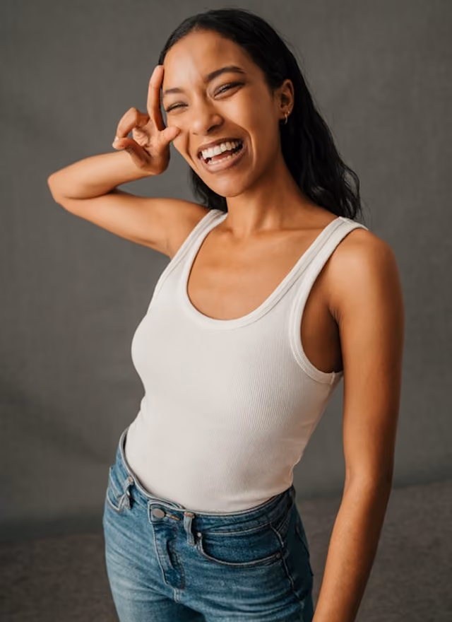 Smiling woman in white tank top and jeans making a peace sign near her eye.