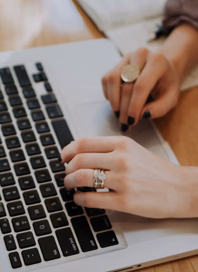 Two hands typing on a laptop keyboard, one hand with a diamond ring and the other with a large ring.