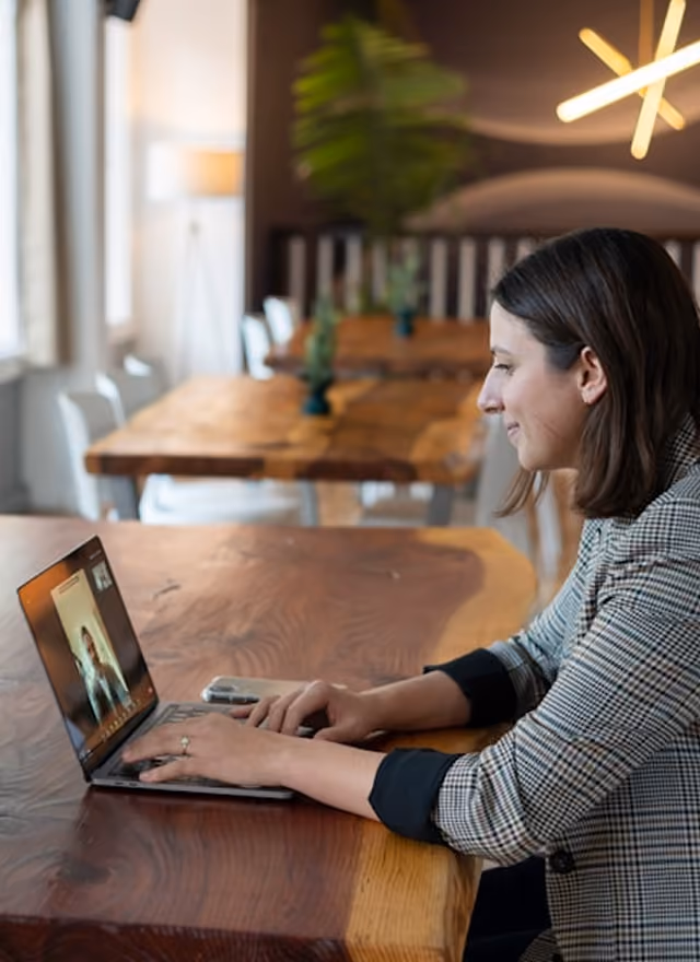 Woman video calling on a laptop at a wooden table in a cozy room with plants and lights.