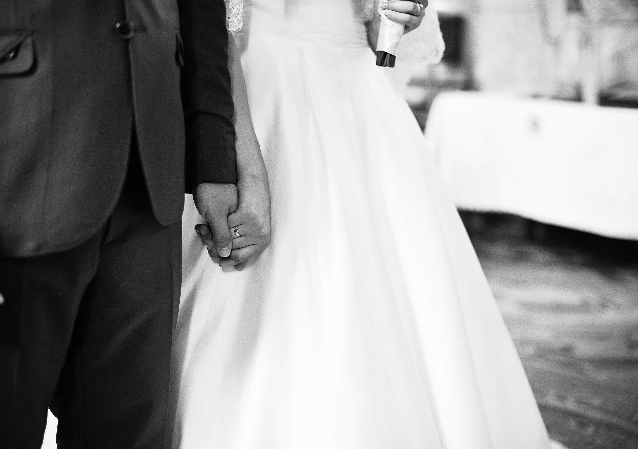 Close-up of a bride and groom holding hands, showing the bride's wedding ring and white dress.