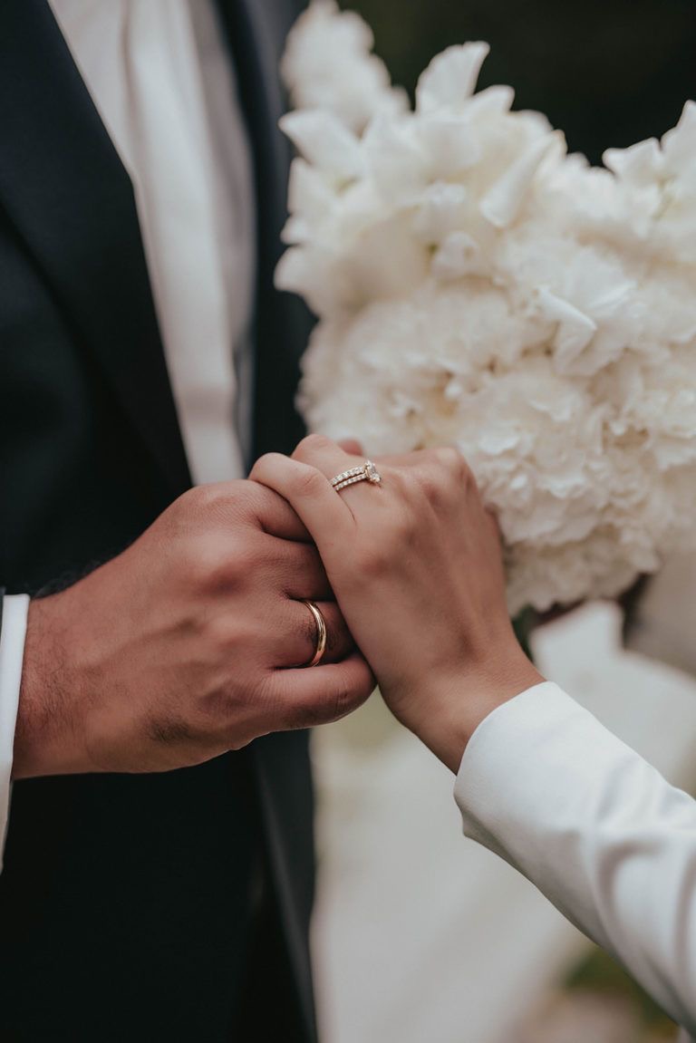 Close-up of a couple holding hands, showing wedding and engagement rings with a white flower bouquet in the background.