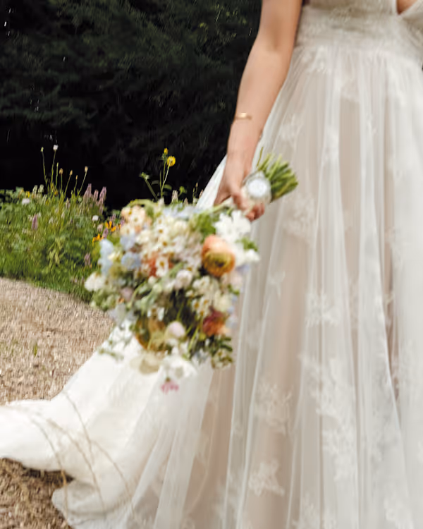 Close-up of a bride holding a colorful flower bouquet wearing a white lace wedding dress outdoors.
