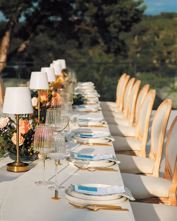 Elegantly set dining table outdoors with white tablecloth, gold utensils, glassware, floral centerpieces, and white cushioned chairs.