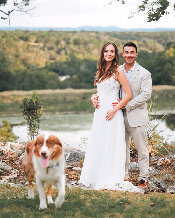 Smiling bride in white dress and groom in light suit standing outdoors near a lake with a brown and white dog in the foreground.